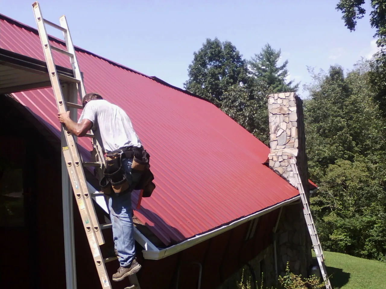 Roofer on a ladder beside a red metal roof and stone chimney — Soffit & Fascia Repair by Roofing Brothers in Gravel Switch